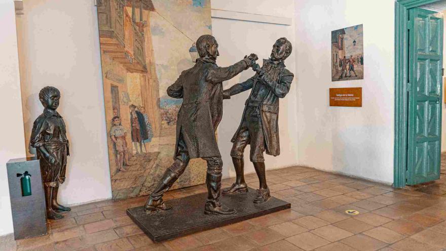 Dos estatuas de bronce de dos hombres que parece que están peleando en la calle, tal y como lo recrea un cuadro de esta y detrás de ellos. Al lado, como otra estatua de bronce, un niño los observa.