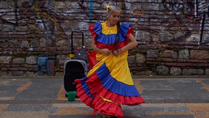 Una mujer adulta y vestida con un vestido con colores de la bandera de Colombia baila en una calle. Hay un bafle que parece que reproduce la música que ella baila.