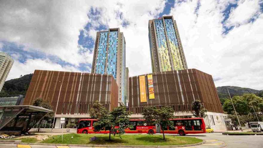  Fachada exterior de un alto edificio en concreto y madera. Detrás hay un par de altas torres residenciales y al frente pasa un bus rojo del sistema TransMilenio. Hay varios árboles alrededor.