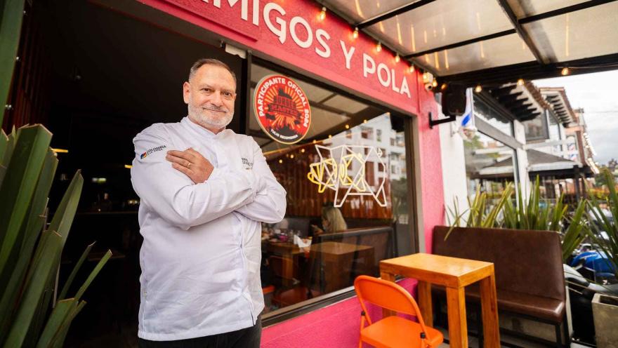 Un chef sonriente con los brazos cruzados, posando frente a la entrada de su restaurante con un diseño vibrante. 