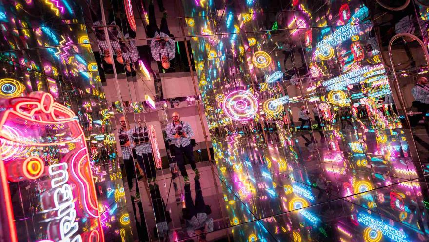 Un hombre parado en una sala llena de espejos y luces de neón vibrantes, creando un efecto infinito y futurista. 