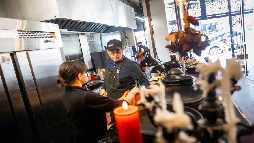 Dos chefs interactuando en una cocina profesional, con utensilios y una vela encendida en primer plano. 
