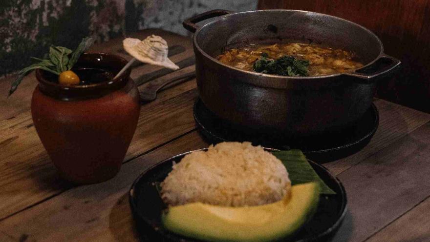  Bodegón con una olla de comida, un plato de arroz con aguacate y una bebida en un recipiente de barro sobre una mesa.