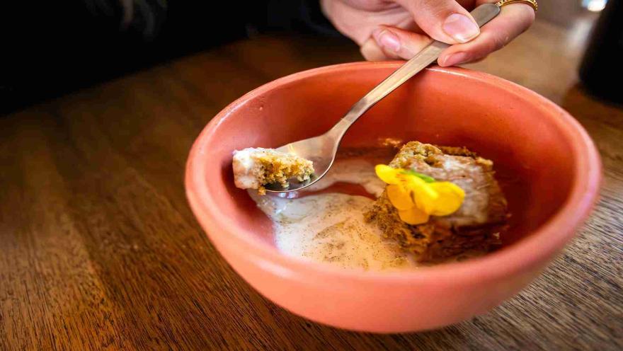 Una cuchara en un tazón de postre con bizcocho, helado y una flor amarilla comestible. 