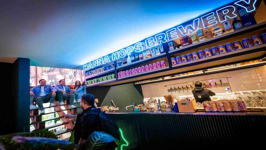 Un bar moderno con letreros de neón, estantes llenos de cervezas y personas disfrutando de la atmósfera vibrante. 
