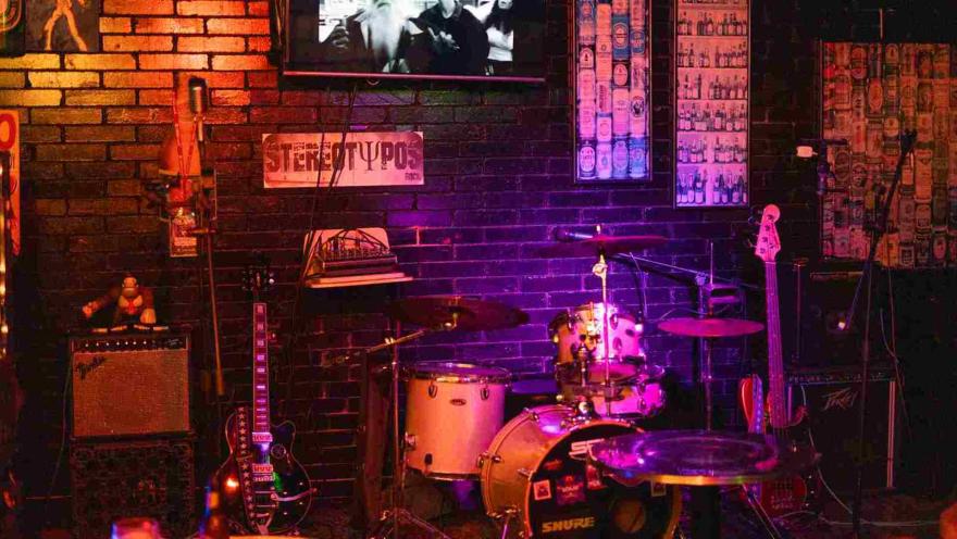 Un escenario de bar con batería, bajo y amplificadores bajo luces de colores, listo para una actuación.