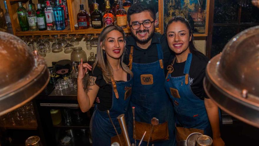 Tres sonrientes bartenders, dos mujeres y un hombre, posan detrás de una barra bien surtida. 