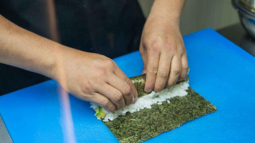 Manos de una persona enrollando un rollo de sushi con arroz y aguacate sobre una tabla de cortar azul.