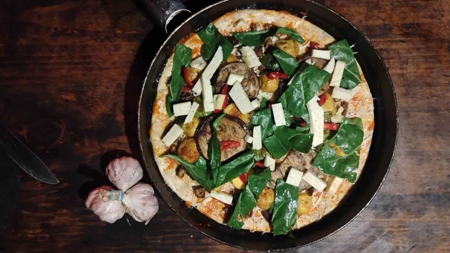 Vista cenital de una pizza con espinacas, berenjena y queso, cocinada en una sartén negra, con una cabeza de ajo al lado sobre la mesa.