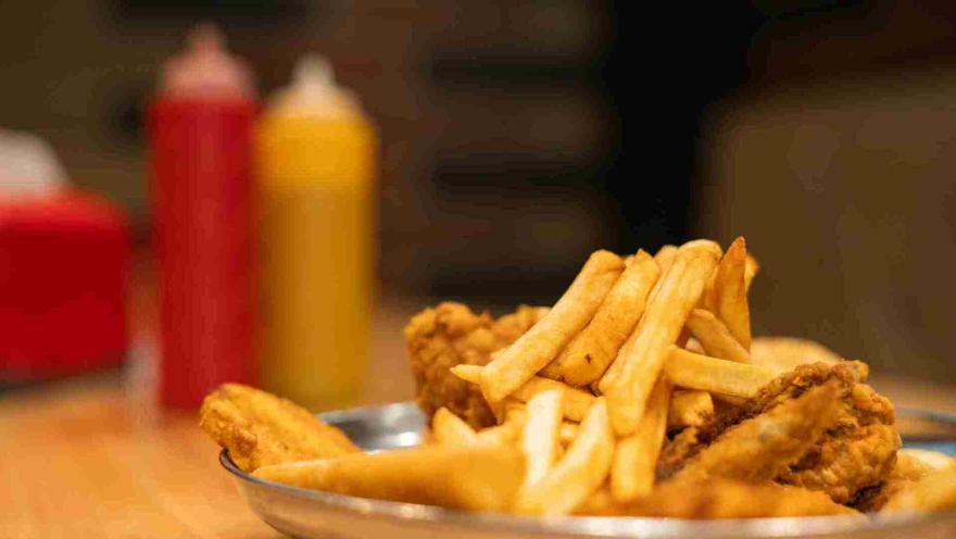 Primer plano lateral de un plato con piezas de pollo frito y papas fritas, con botellas de salsa desenfocadas al fondo.