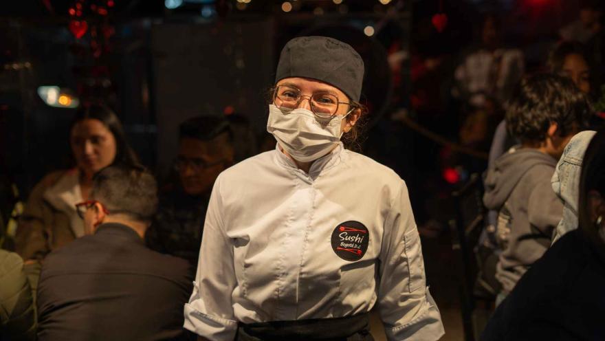 Chef de sushi mujer con uniforme, gafas y mascarilla, mirando a la cámara y sonriendo en un ambiente nocturno.