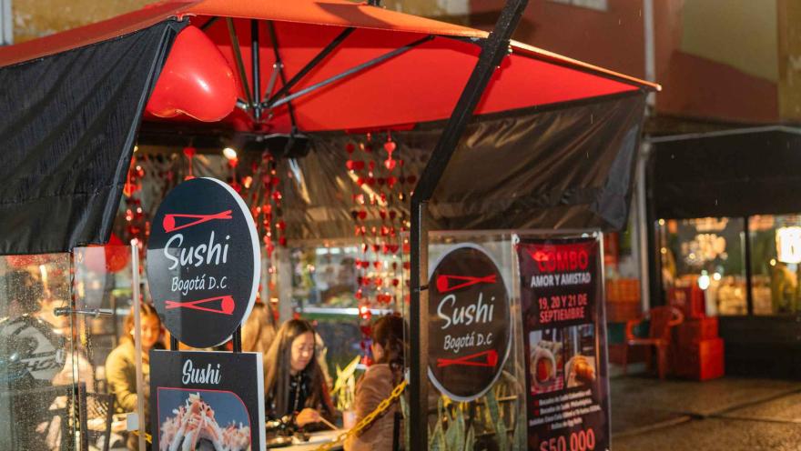 Puesto de comida al aire libre llamado "Sushi Bogotá D.C." con carteles de menú y clientes sentados.