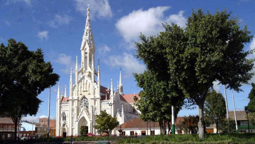 Plano general de la iglesia de estilo gótico con varias puntas hacia el cielo y de color crema. Hay un parque con árboles alrededor.