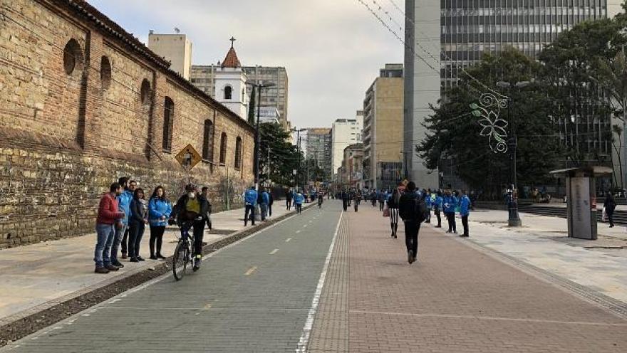 En el centro, la Carrera Séptima luce organizada y abierta, con un carril para bicicletas y amplios espacios para peatones. Personas caminando, conversando y un ciclista en movimiento reflejan un ambiente tranquilo y dinámico, enmarcado por edificios modernos y árboles que aportan equilibrio al paisaje urbano.