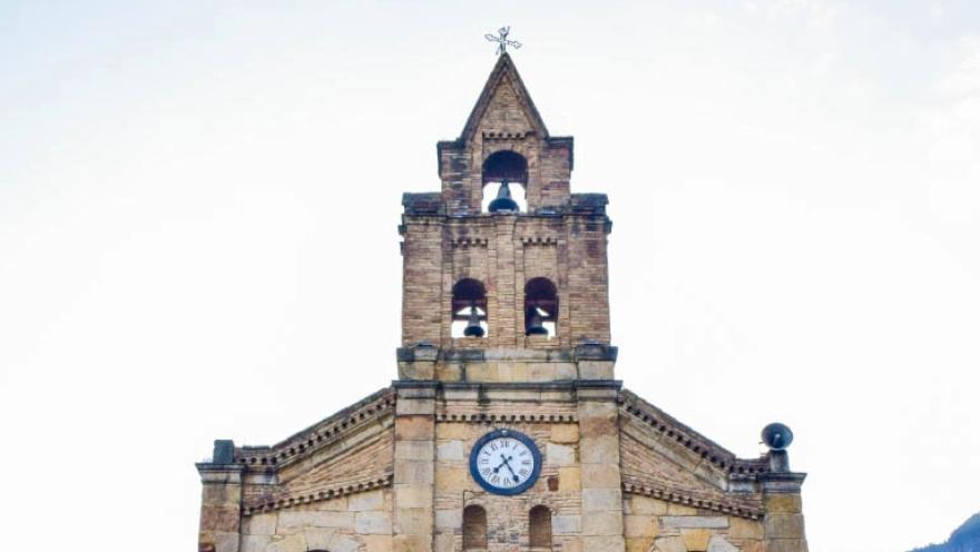  Primer plano de la parte alta de la iglesia en piedra en la que sobresale la torre con el campanario y el reloj.