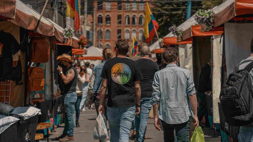 Un grupo de personas camina por entre una calle rodeada por toldos de comercio. Alrededor hay varias banderas de Colombia.