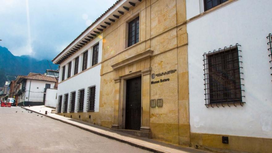  Fachada del Museo Botero vista de lado. Es una casa de dos pisos en piedra y concreto pintado de blanco. En la entrada se alcanza a leer una leyenda que dice: "Museo Botero"