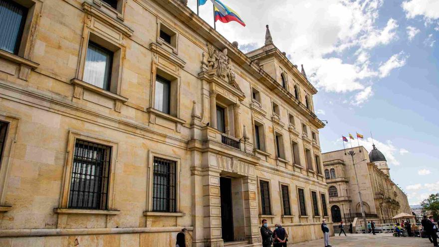  Fachada del edificio en piedra visto de lado y en el que sobresale una bandera de Colombia y otra de otro lugar, en el techo. Hay varias personas alrededor. 