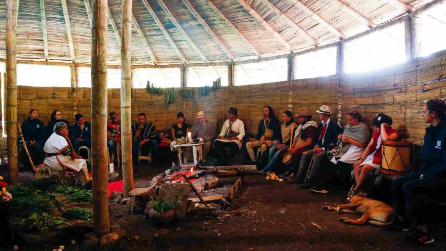 Un grupo diverso de personas reunidas dentro de una estructura circular que parece una choza tradicional. El techo está hecho de madera y paja, sostenido por pilares de madera. Las paredes están hechas de bambú y hay ventanas rectangulares a lo largo de la parte superior de la pared que dejan entrar la luz natural.