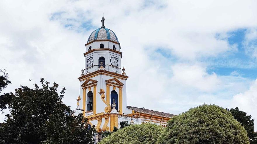  Vista de la torre principal de una iglesia en la que se observa un campanario y varios relojes. En la punta de la figura en forma de óvalo hay una cruz metálica.