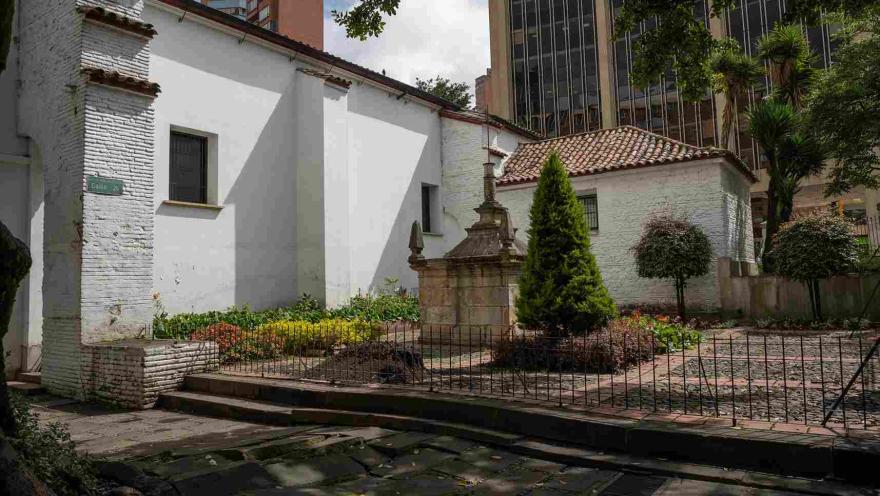 Parte exterior de la iglesia pintada de blanco. Hay un jardín con un pino y una estructura en piedra.