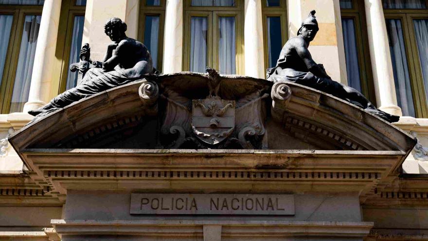 Primer plano de fachada de edificio en la que se observan dos figuras en piedra con forma de soldados sentadas en el techo, el escudo de Colombia, también en piedra, y una leyenda con la frase “Policía Nacional”.