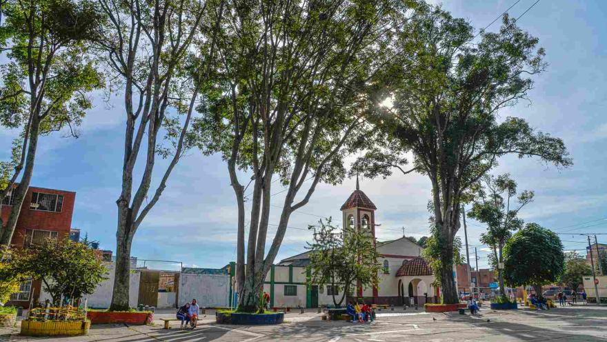 Una plaza tranquila de barrio, rodeada de altos árboles que filtran la luz del sol y proyectan sombras sobre el suelo. En el centro se alza una pequeña iglesia de fachada blanca y campanario con tejas rojas, que aporta un aire tradicional al entorno.