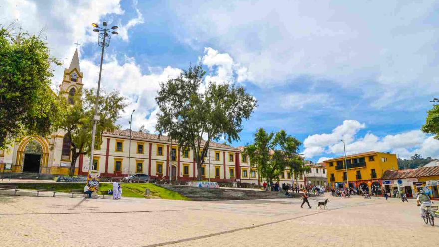 Una plaza de pueblo con una iglesia, edificios coloridos y personas disfrutando del día. En la imagen, se ve una amplia plaza pavimentada con adoquines de color claro. En el fondo, se encuentra una iglesia con una torre alta y puntiaguda, pintada de blanco y amarillo. A la derecha de la iglesia, hay un edificio de dos pisos con paredes blancas y detalles en rojo y amarillo alrededor de las ventanas. Un gran árbol está frente al edificio, proporcionando sombra.