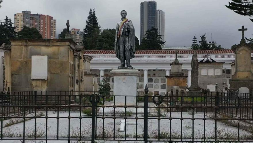 Estatua en piedra en mausoleo, parece ser del general Santander con una cinta con los colores de la bandera de Colombia. Al lado hay varios mausoleos de piedra. Al fondo, arriba, a lo lejos se observan edificios.