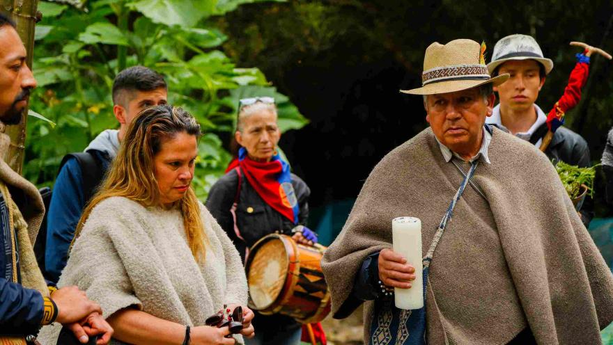 Un grupo de personas, algunas de las cuales parecen estar vestidas con atuendos tradicionales. En primer plano, un hombre lleva un sombrero de paja tejido y un poncho marrón; está sosteniendo una vela blanca. A su izquierda, una mujer con un suéter color crema está mirando un objeto pequeño en sus manos. Detrás de ella, una mujer sostiene un tambor, y lleva un pañuelo rojo, azul y amarillo alrededor del cuello. En segundo plano, se ven otras personas y vegetación. La escena parece tener lugar al aire libre.