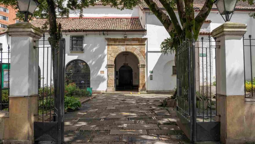  Parte exterior de la puerta principal de la iglesia en la que se ve una reja que da paso al antejardín y el arco en piedra de la puerta al fondo.