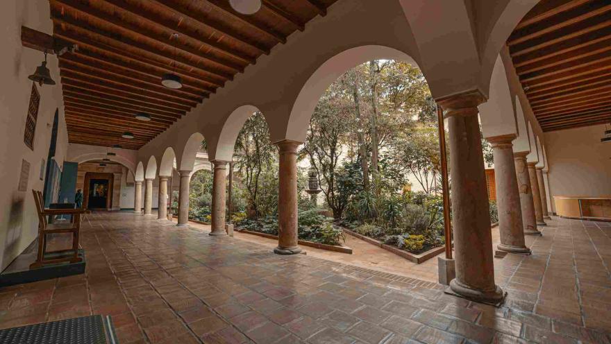 Un primer piso en el que hay un largo pasillo en ángulo con unas columnas que forman arcos y sostienen un segundo piso. Al fondo se observa un patio con un jardín.
