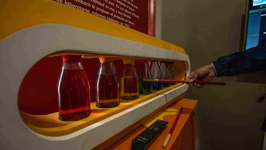 Una exposición de botellas de vidrio con un líquido de color específico en cada una. Hay una mano que parece estar señalando algo.