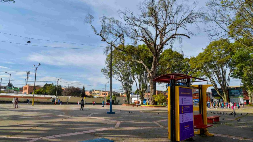 Una amplia plaza pública en un día despejado, con un ambiente tranquilo y cotidiano. El espacio está pavimentado con grandes losas de concreto, marcadas por líneas diagonales de color rosado pálido que crean un diseño geométrico en el suelo. En el primer plano, a la derecha, destaca una estructura informativa de color amarillo y morado con un techo rojo, acompañada de un banco de madera rojiza. A la izquierda, se observa un banco largo y sencillo de color azul intenso.