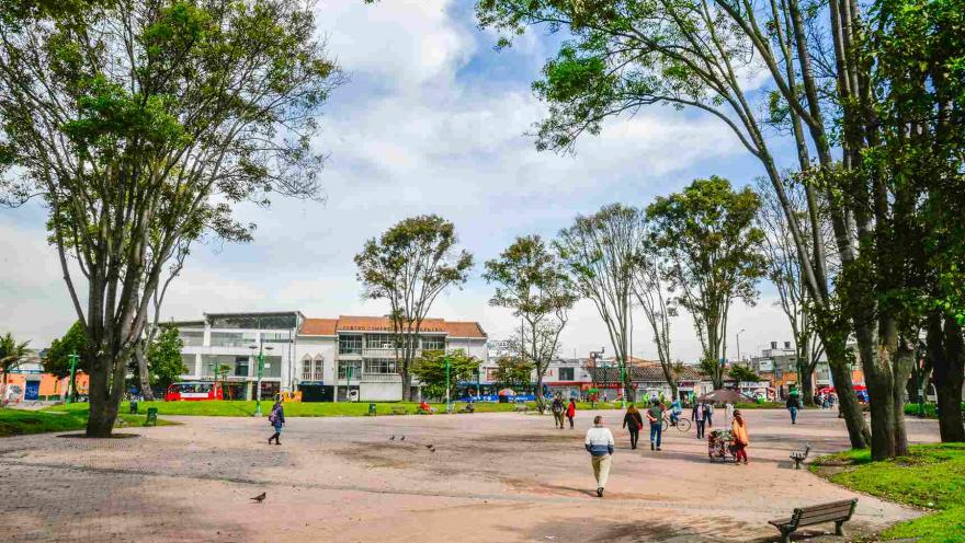 Plano general de la plazoleta central donde hay varios árboles altos y personas que caminan alrededor.