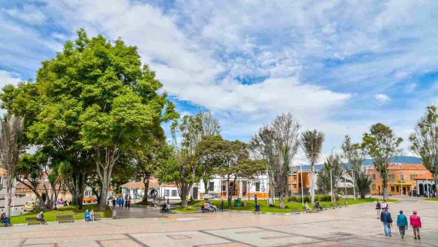 Una plaza de pueblo con edificios coloridos y personas disfrutando del día. En la imagen, se ve una amplia plaza pavimentada con adoquines de color claro. 