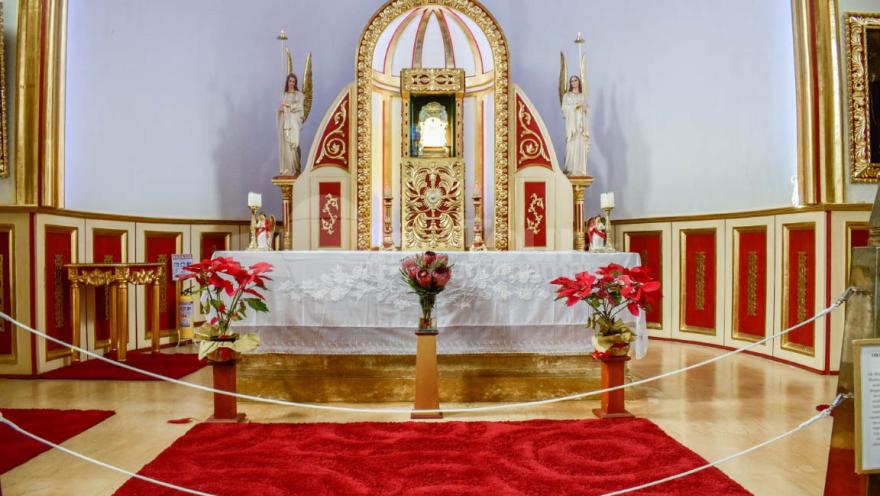 Primer plano interior de la parroquia en la que se ve el altar decorado con tres arreglos florales y varios tapetes de color rojo.
