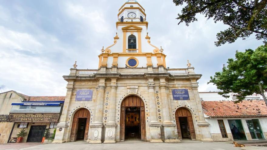  Fachada de la Parroquia de Santa Lucía, vista de frente y desde abajo, en la que destacan sus tres grandes y ovaladas puertas de madera abiertas, la torre principal y un reloj.