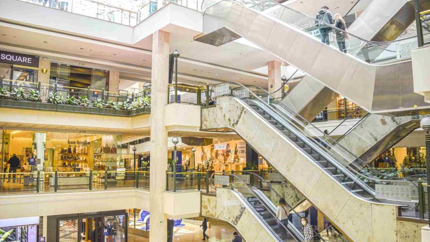 Interior de centro comercial en el que sobresalen escaleras eléctricas, locales comerciales en varios pisos y algunas personas alrededor.