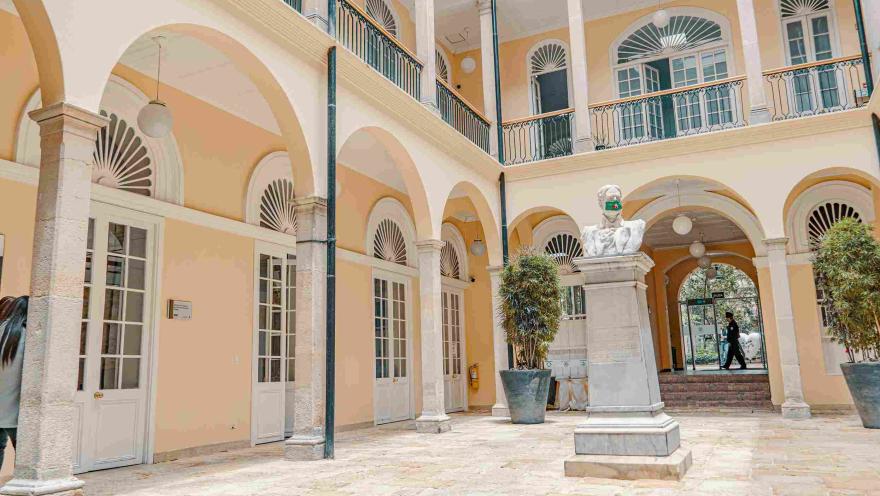  Primer plano del interior del palacio en una especie de patio en el que se observa una estructura de dos pisos color crema, el segundo con bellos balcones y en el primero varias columnas en forma de arco. En el centro del lugar hay una estatua, parece que de un héroe patrio.