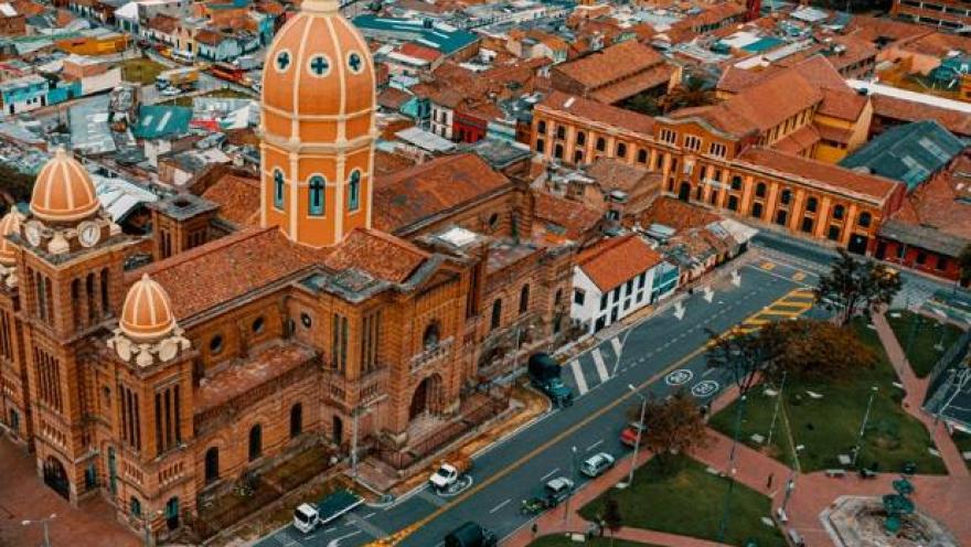 Panorámica de una iglesia vista desde arriba, como con un dron. Sobresalen los techos de ladrillo y una torre alta en forma ovalada. Se observa un parque al frente de la iglesia.