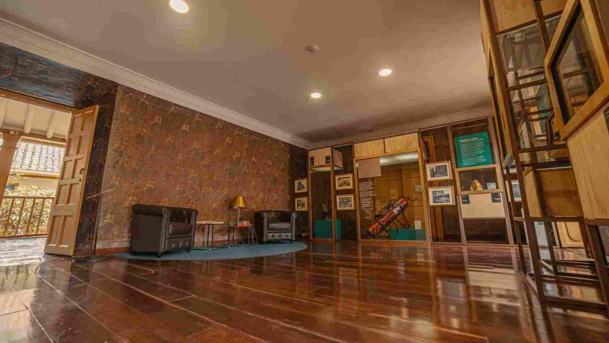Salón interior con un piso en madera brillante y en el que se observan algunos estantes en madera con diversos artículos, entre ellos un extintor gigante. En el salón hay dos poltronas de cuero, separadas por dos mesas pequeñas; en una de ellas hay una lámpara metálica dorada.