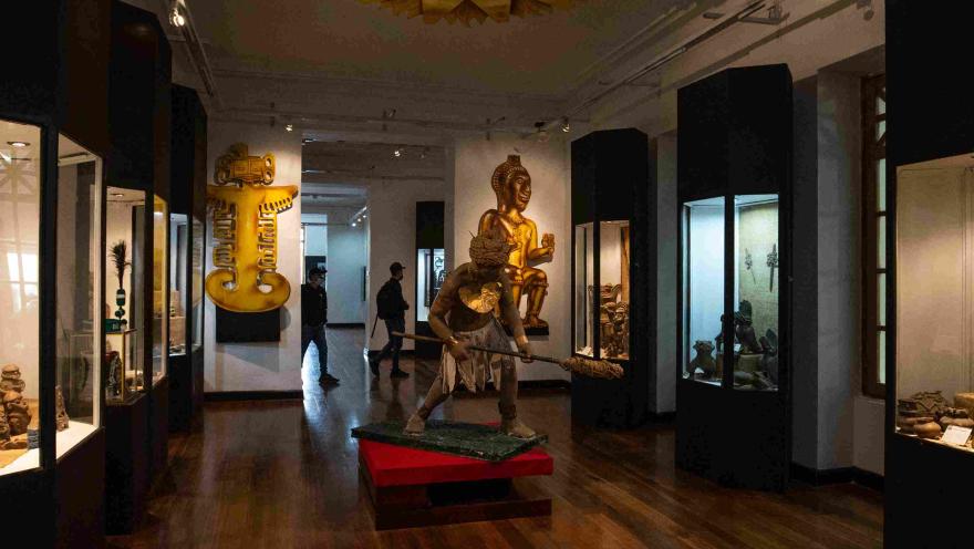 Interior de museo con varias galerías en vidrio que contienen diferentes piezas que parecen ser precolombinas, por los objetos decorativos de gran tamaño alrededor, tales como figuras indígenas.