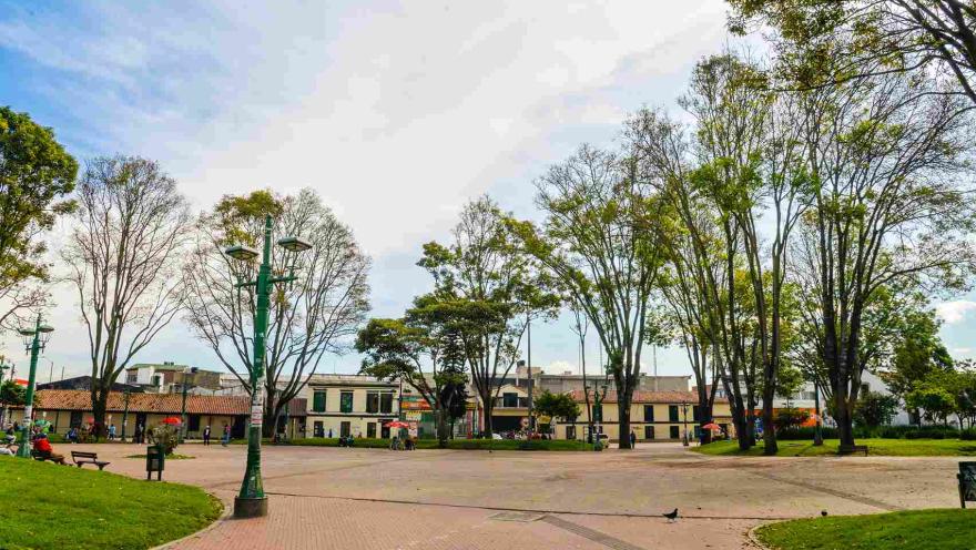 Plano general de la plazoleta central donde hay varios árboles altos, faroles y varias casas al fondo.
