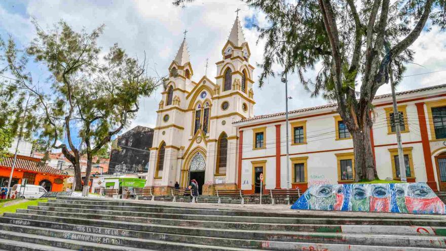  Vista inspiradora de una iglesia histórica con dos campanarios en Bogotá, rodeada de frondosos árboles y vibrante arte callejero en las escaleras, que simboliza el patrimonio cultural y el espíritu comunitario.