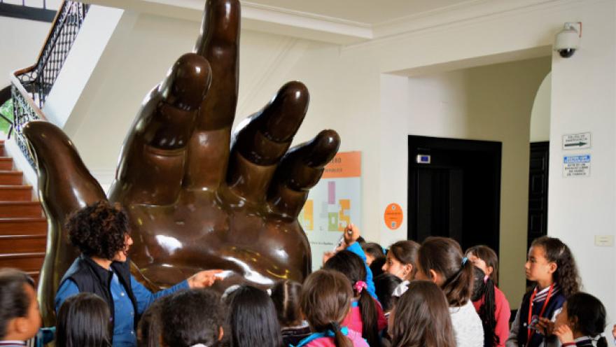 Una escultura en forma de mano gigante y de color café rodeada por niños y una mujer adulta que parece ser una guía o una profesora.