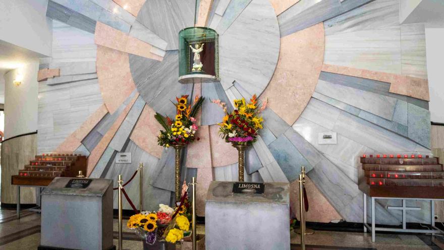 Plano general del interior de la capilla donde en una urna de cristal está el Divino Niño.  Hay tres arreglos florales alrededor.