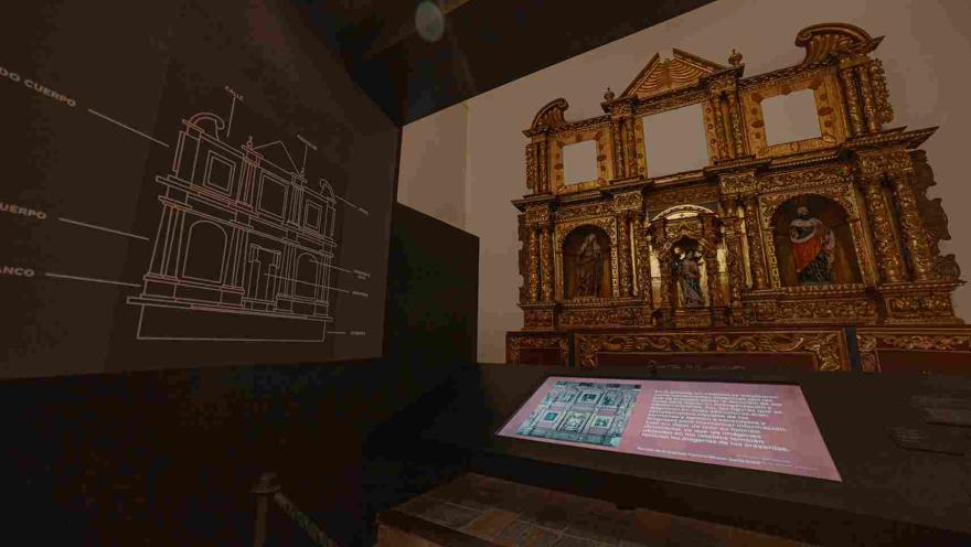 Un pedazo grande de retablo dorado está exhibido en una pared. Hay tres imágenes de santos que lo acompañan. Hay una foto de un plano de una casa que acompaña la escena.