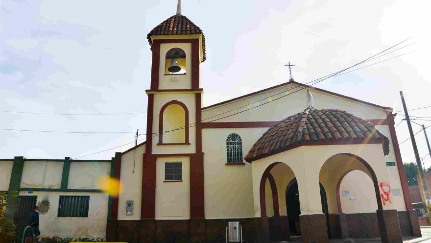  Una iglesia de estilo colonial con una torre campanario prominente. La estructura principal está pintada en tonos crema claro con detalles en rojo ladrillo, creando un contraste visual llamativo. El techo es de tejas rojas, y sobre el edificio principal se alza una cruz.