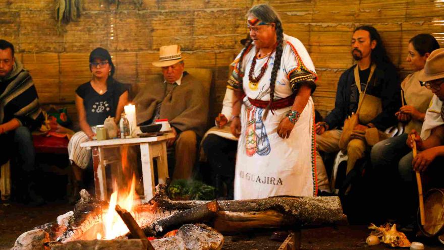 Una escena en el interior de una estructura rústica con paredes de bambú. Un grupo de personas se encuentran sentadas a lo largo de las paredes. Destaca una figura femenina de pie, vestida con un atuendo tradicional blanco adornado con símbolos y la palabra "Guajira". Lleva el pelo en trenzas y luce un tocado y collares coloridos. Delante de la figura, hay una fogata encendida con leños gruesos y rocas alrededor, que ilumina la escena con llamas y luz cálida. 